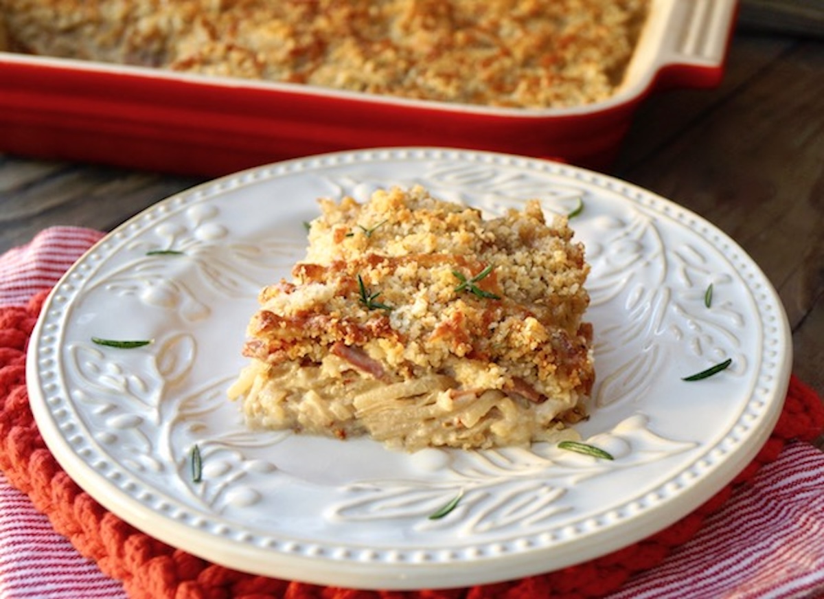 One square of Funeral potatoes (Rosemary Prosciutto Cheesy Idaho® Potatoes) on a white plate with fresh rosemary.