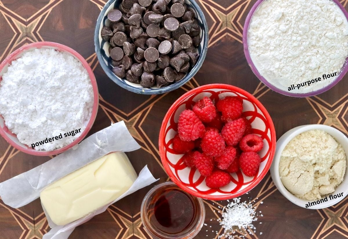 Raspberry-Chocolate-Chip-Cookies Ingredients for Raspberry chocolate chip cookies on a dark wood cutting board, including: chocolate chips, fresh raspberries, butter, vanilla, salt, powdered sugar, flour and almond meal.