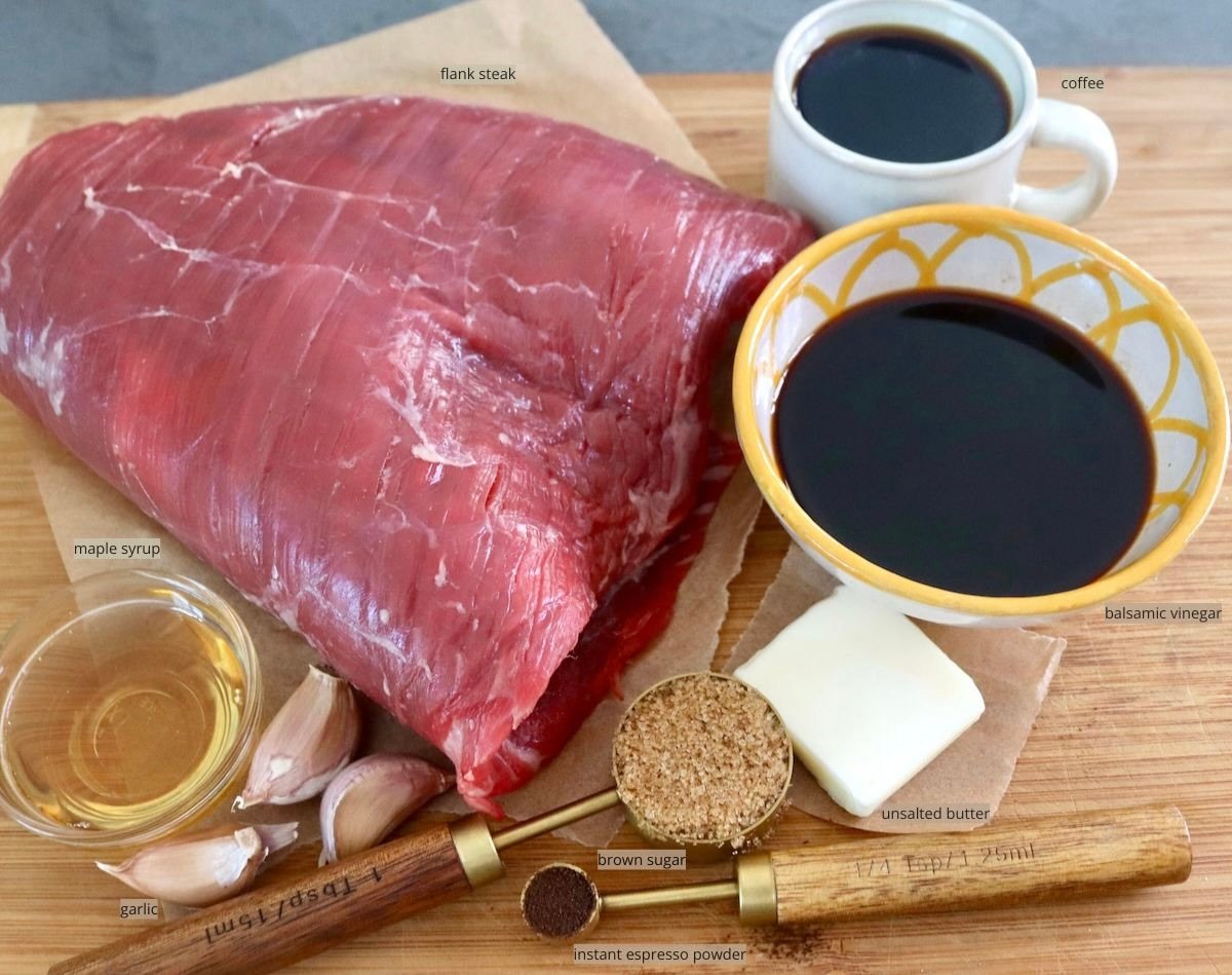 Raw fank steak, bowl of balsamic vinegar, pat of butter, cup of coffee, garlic cloves, and maple syrup, on a cutting board.
