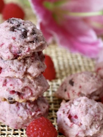 Stack of 4 raspberry chocolate chip cookies with a pink lily flower behind them, with a few fresh bright red raspberries around them.