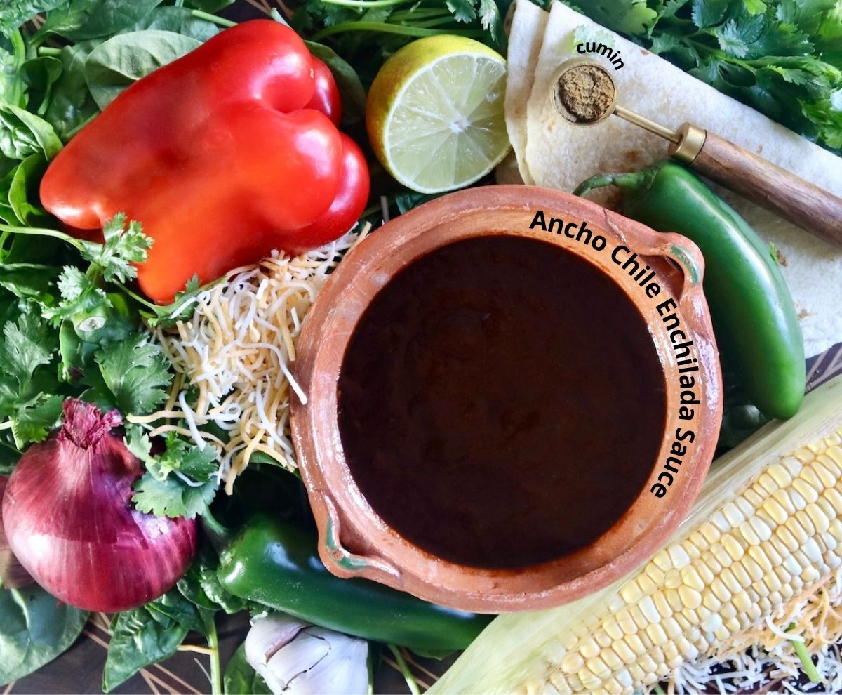 Ingredients for vegetarian enchiladas including red pepper, jalapenos, lime, bowl of ancho chile sauce, red onion, grated cheese, spinach and cilantro.