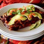 Two enchiladas on a white plate with thinly sliced avocado and diced tomato.