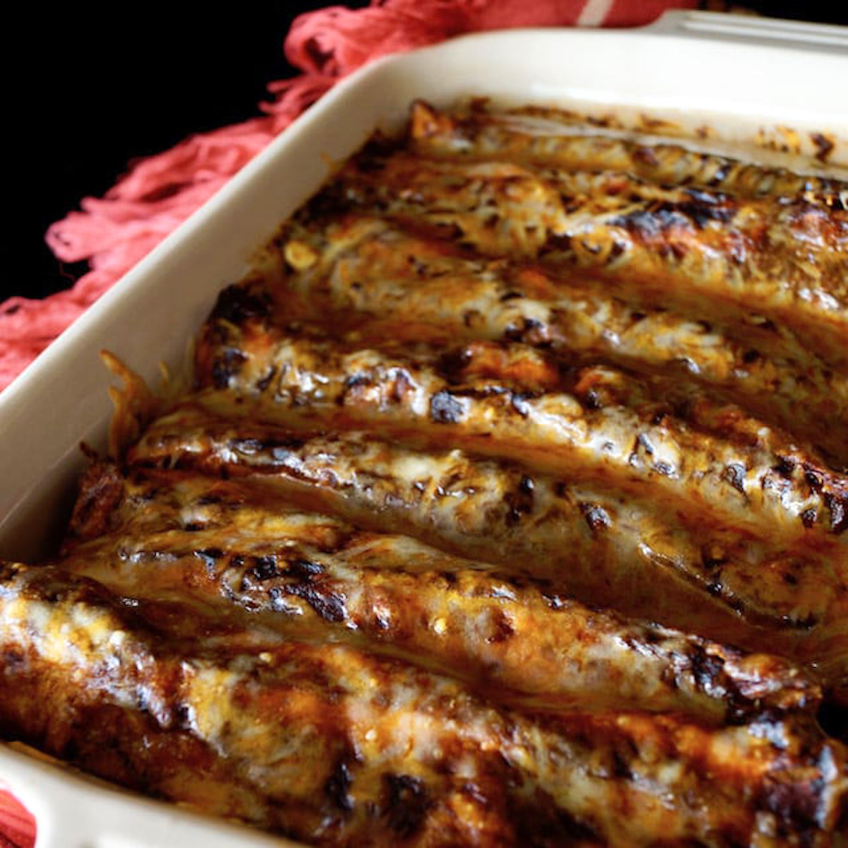 Ceramic baking dish with several baked vegetarian enchiladas with sauce and cheese, on a red cloth napling with fringe.