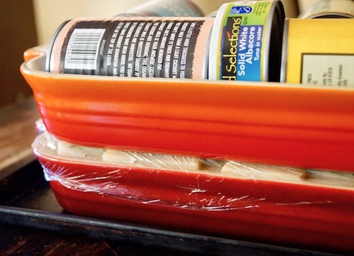 Two red ceramic casserole dishes on top of each other with cans of food in the one on top, to weigh down the potato gratin beneath it.