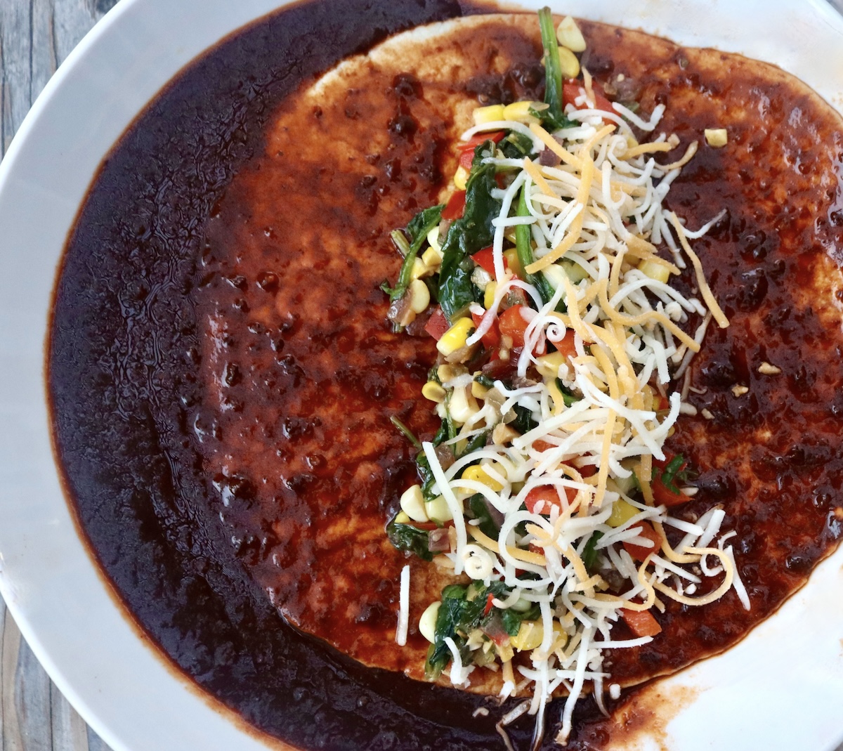 One enchilada-sauced tortilla with a vegetable filling and shredded cheese in the middle, ready to be rolled.