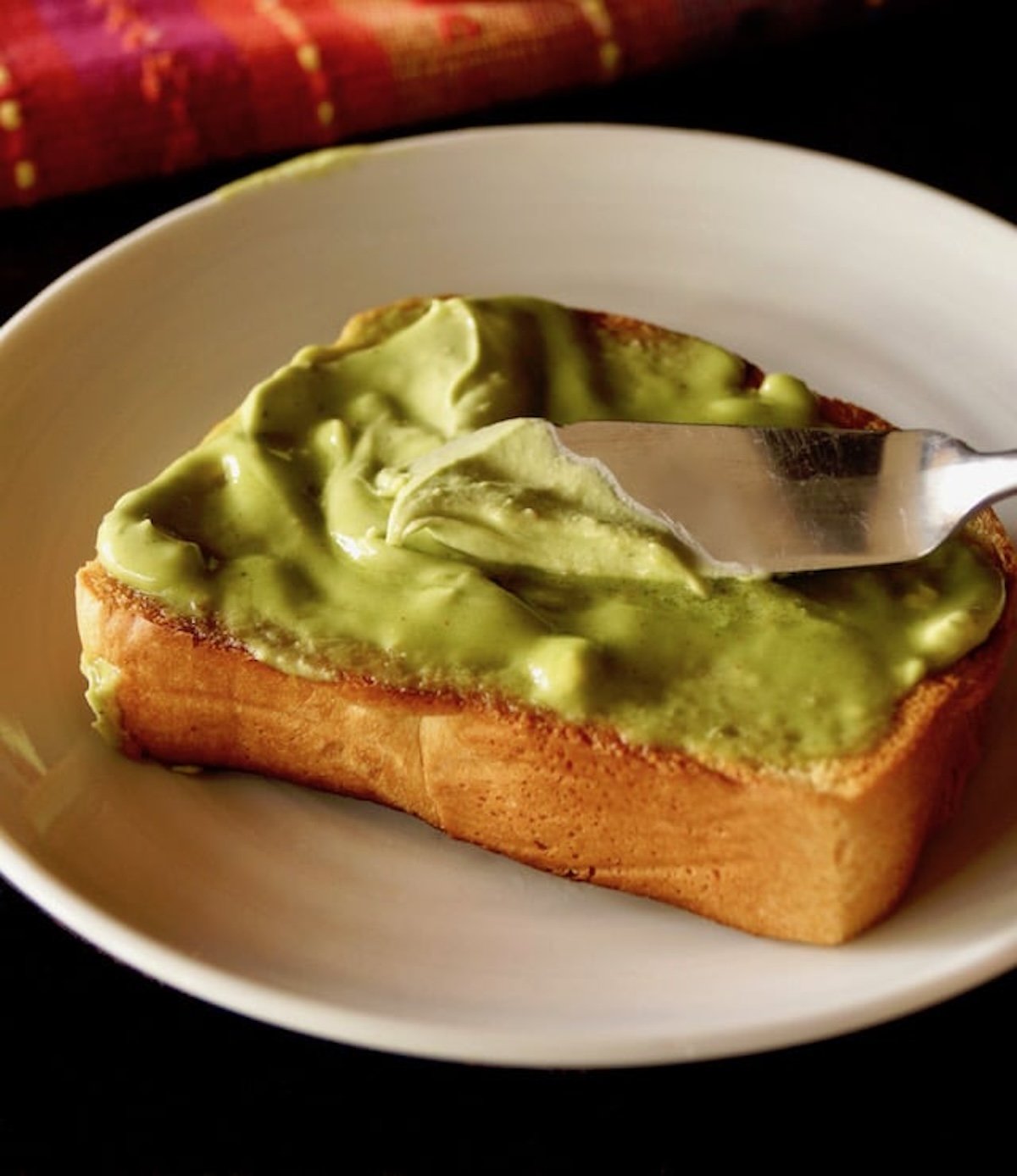 Soft Avocado Butter spread on a thick slice of toast on a white plate with a small spreader.