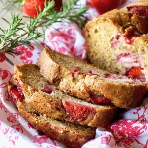 Sliced strawberry loaf cake on a red and white cloth with sprigs of rosemary and whole strawberries.