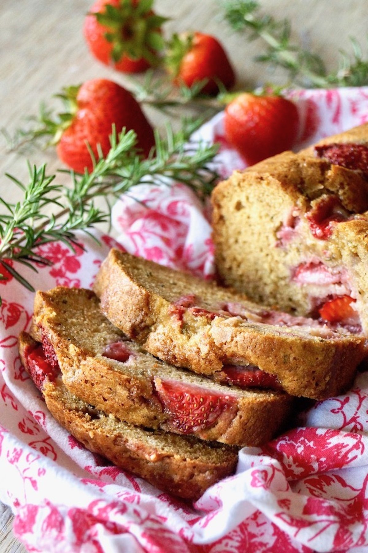 Strawberry-Rosemary-Cake-Recipe Sliced strawberry loaf cake on a red and white cloth with sprigs of rosemary and whole strawberries.