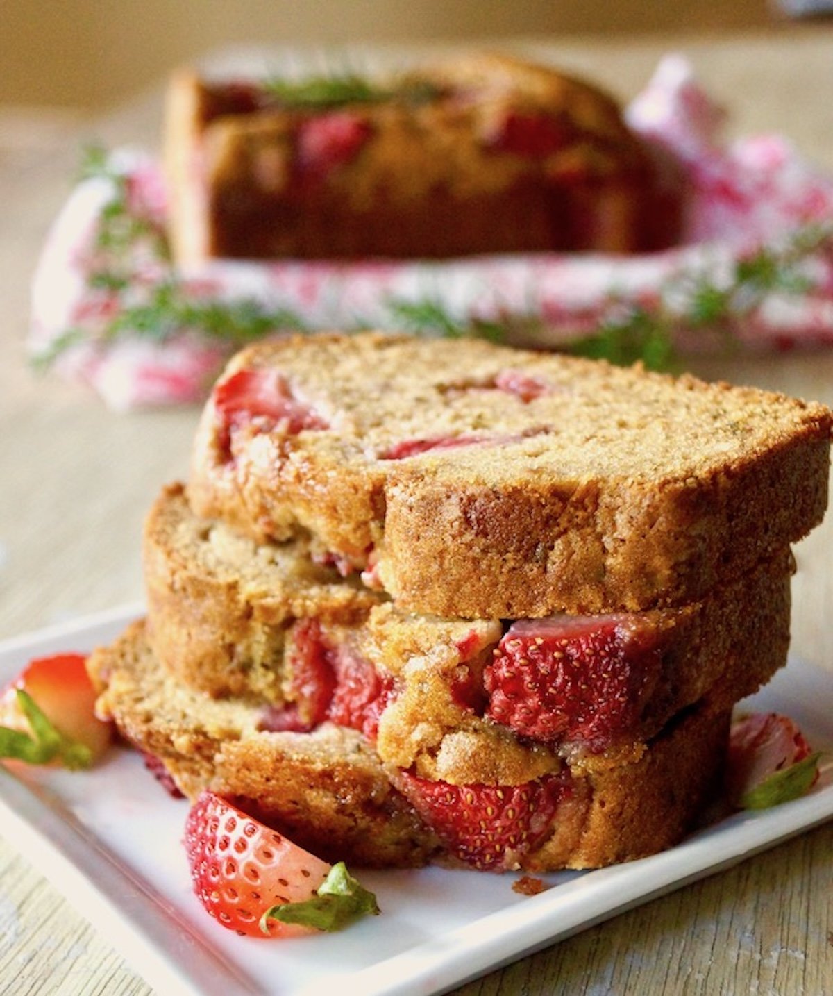 A few stacked slices of strawberry loaf cake on a small white plate with sprigs of rosemary and whole strawberries.