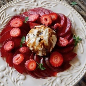 Sliced Elephant Heart plums with strawberry slices on a white textured plate with balsamic drizzle and a few oregano sprigs.