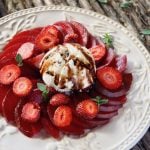 Sliced Elephant Heart plums with strawberry slices on a white textured plate with balsamic drizzle and a few oregano sprigs.