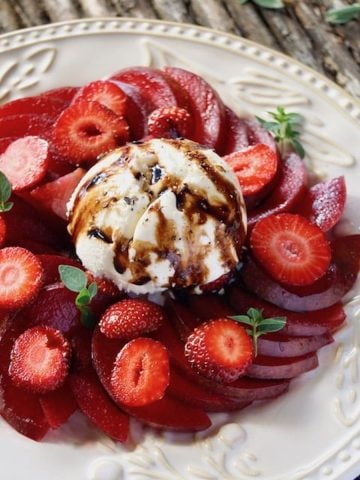 Sliced Elephant Heart plums with strawberry slices on a white textured plate with balsamic drizzle and a few oregano sprigs.