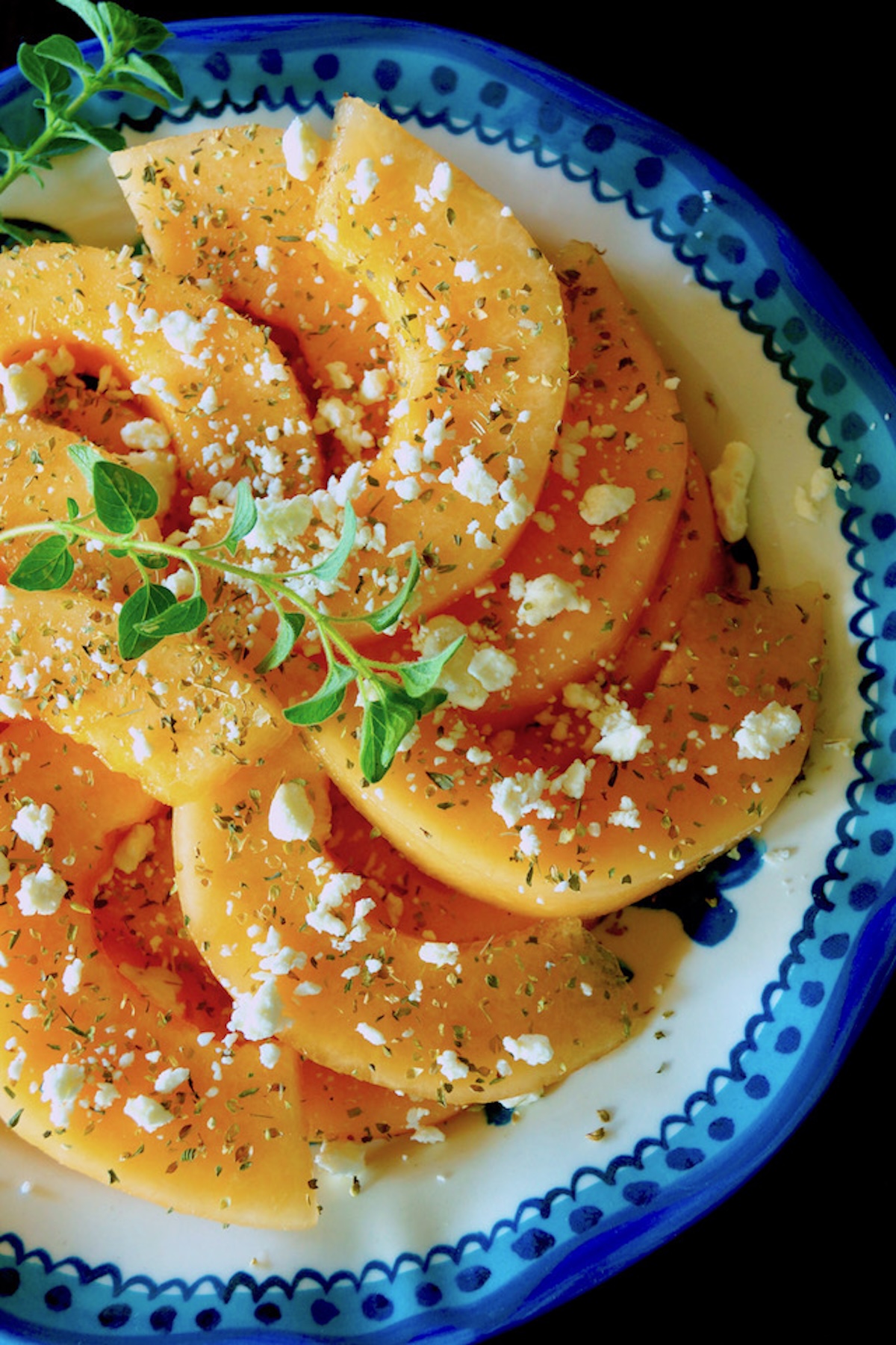 Blue-rimmed plate with sliced orange melon sprinkled with fresh and dried oregano and crumbled Feta.