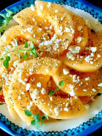 Blue-rimmed plate with sliced orange melon sprinkled with fresh and dried oregano and crumbled Feta.