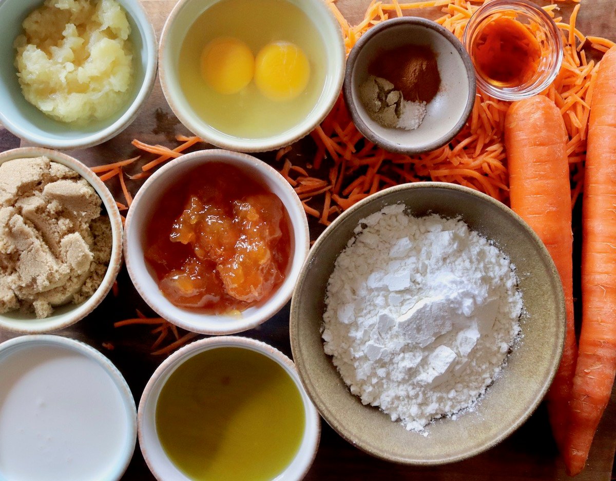 Ingredients for carrot cake in bowls including eggs, flour, apricot preserves, olive oil, spices, sugar and pineapple.