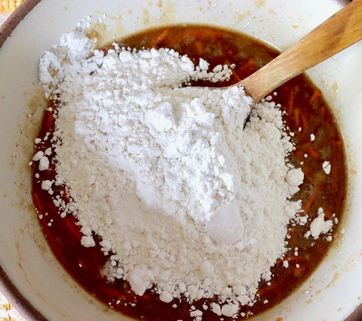 Carrot cake batter with flour poured on top with a wooden spoon.