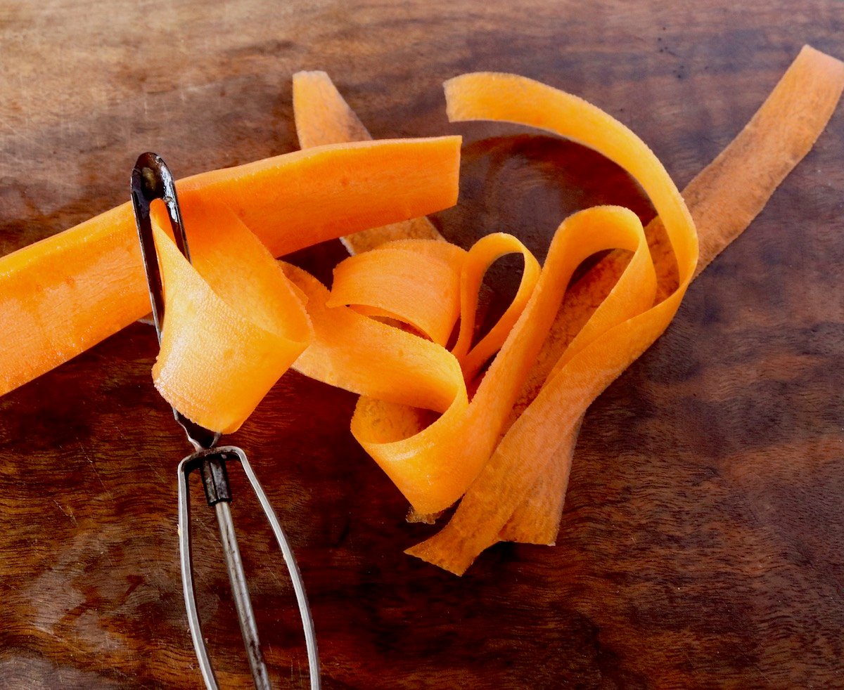 Carrot being peeled into ribbons on a dark wood cutting board.