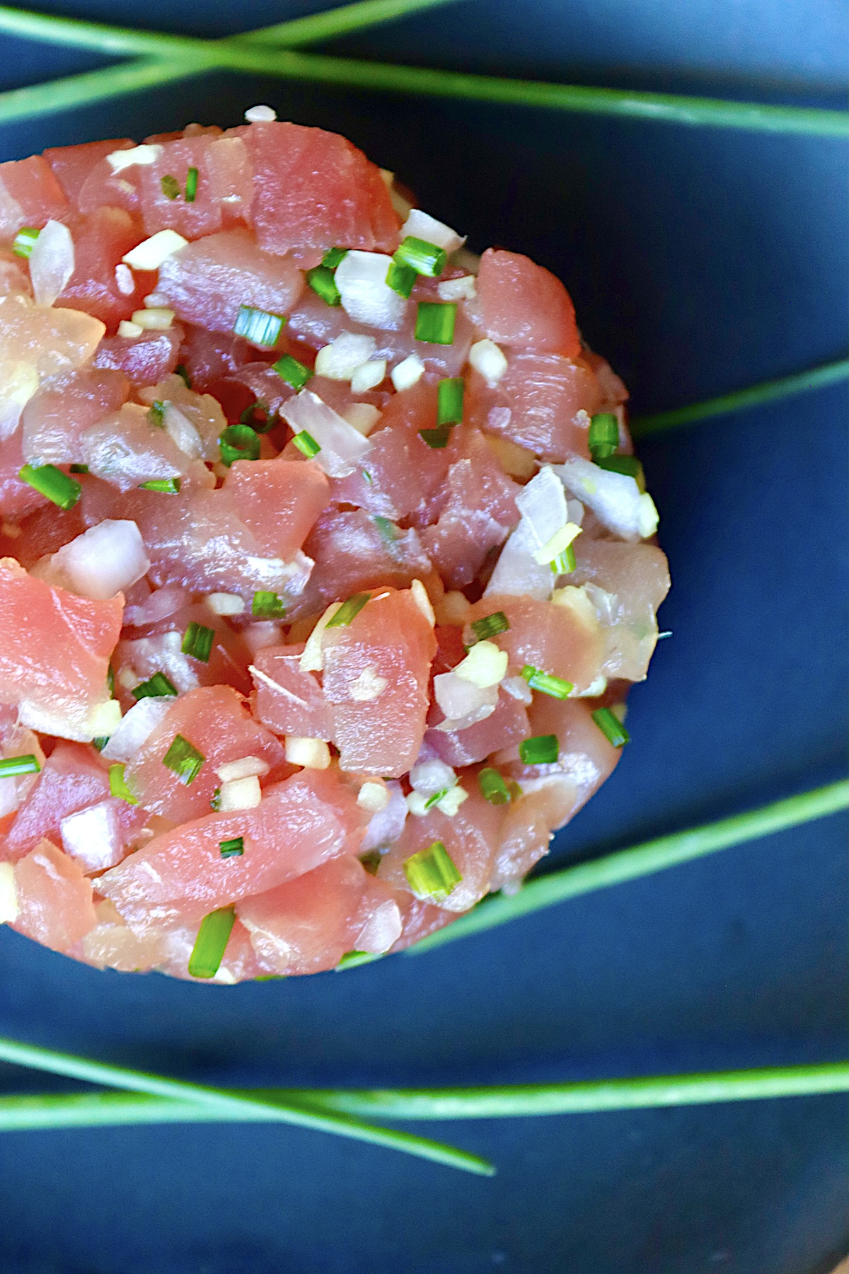 Ahi-Tuna-Tartare-with-Ginger Top view of a round mound of tuna tartare with bits of ginger and onion, on a dark blue ceramic plate.
