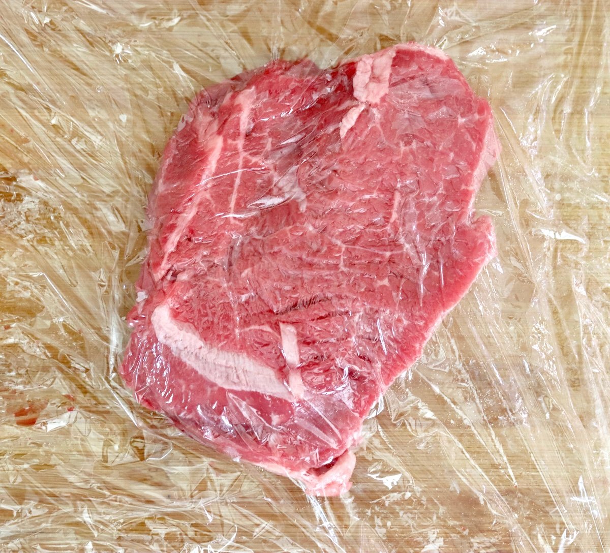 One serloin steak between two sheets of plastic wrap to be pounded thinly.