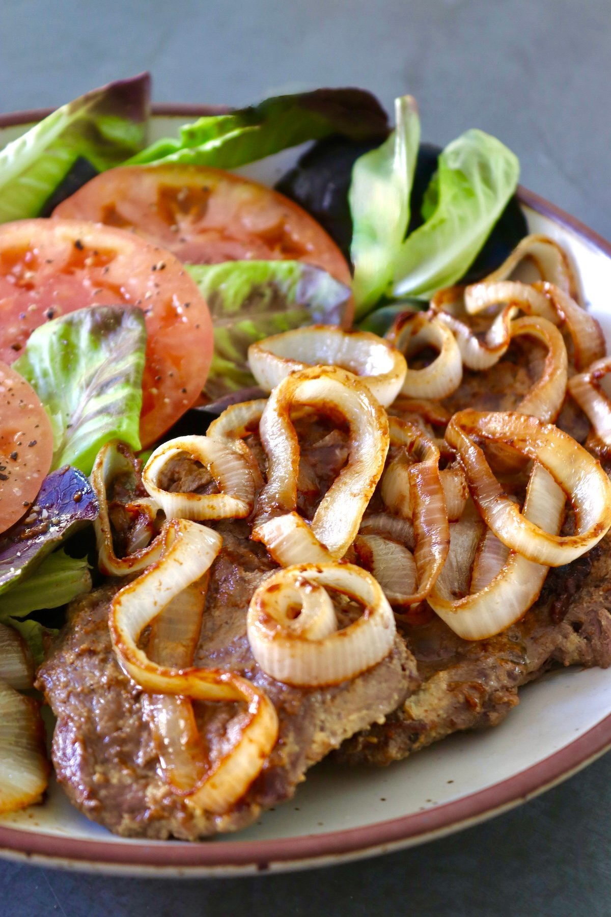 Close up shot of ceramic cream-colored round serving platter with steak and caramelized onion and tomato and lettuce next to it.