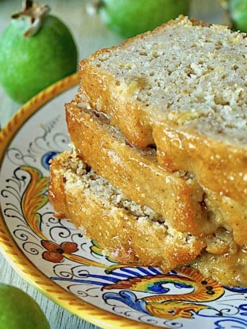 Three slices of Glazed Pineapple Guava Bread on an Italian ceramic, gold-rimmed plate, surrounded by fresh green guavas.