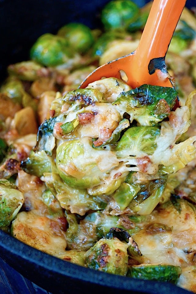 Cheesy Pancetta Brussels Sprouts Bake being spooned out of cast iron skillet with an orange spoon.