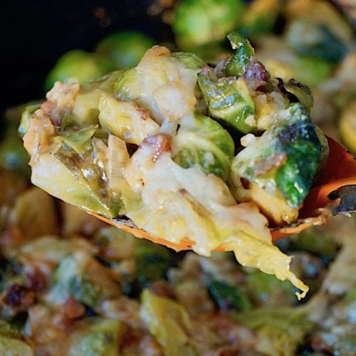 Cheesy Pancetta Brussels Sprouts Bake with a spoonful held above a cast iron skillet with an orange spoon.