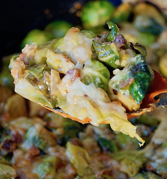 Cheesy Pancetta Brussels Sprouts Bake with a spoonful held above a cast iron skillet with an orange spoon.