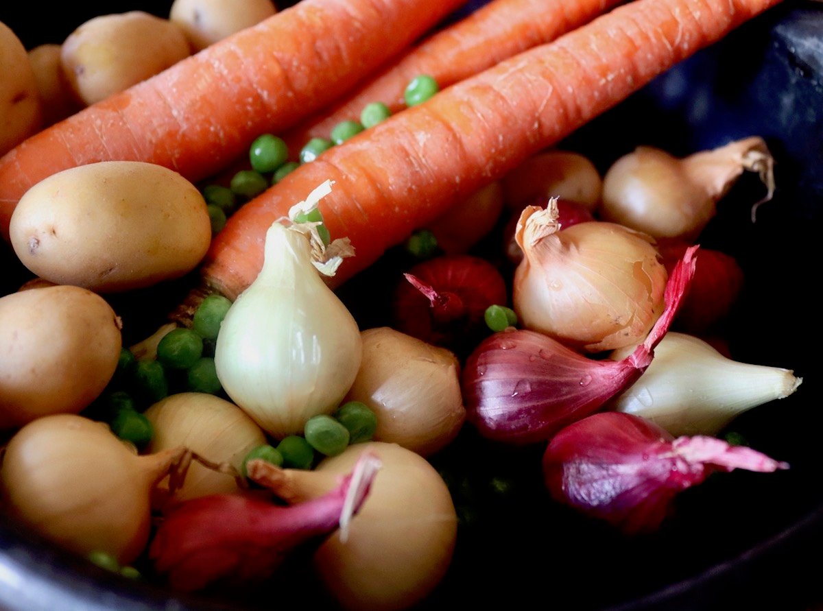 Red and yellow pearl onions with carrots, tiny potatoes and peas in a black bowl. 
