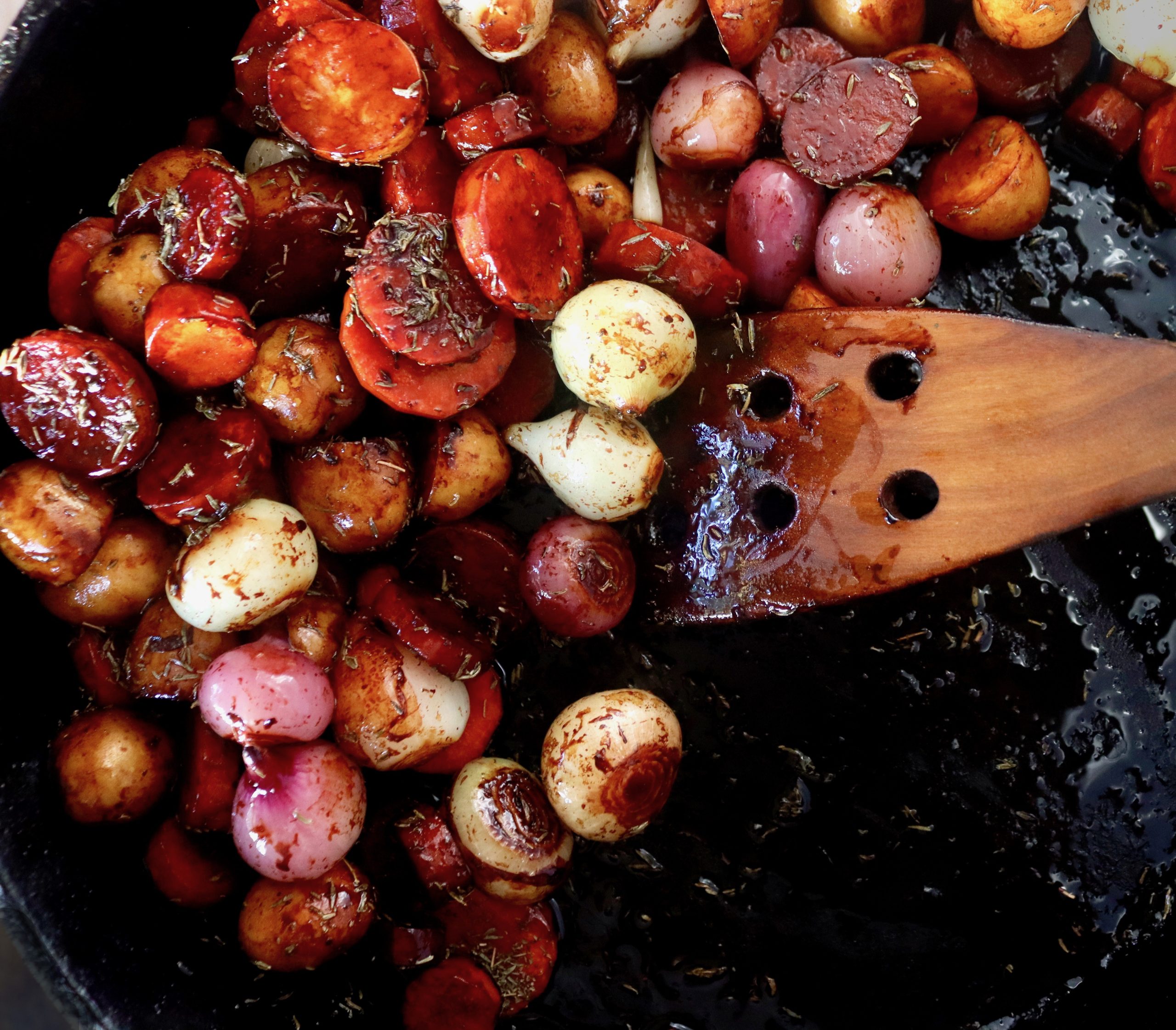 Caramelized carrot slices with potatoes and pearl onions in cast iron skillet with wooden spatula.