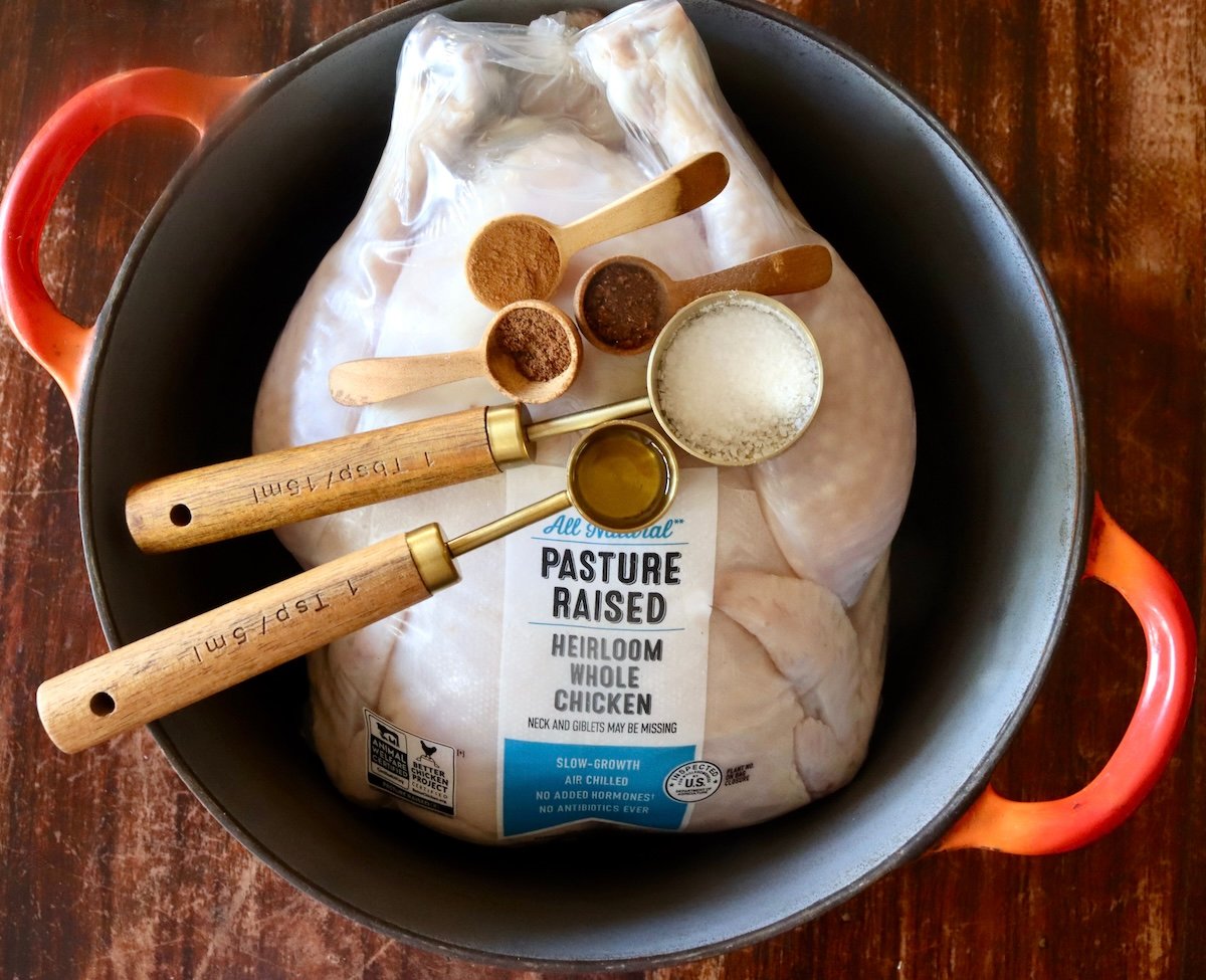 Whole chicken in its packaging, in a Dutch Oven with a few spices in spoons on top.