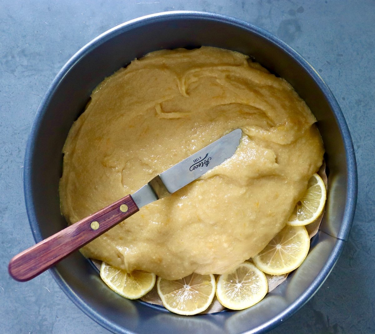 Cake pan lined with lemon slices and lemon cake batter being spread on top.