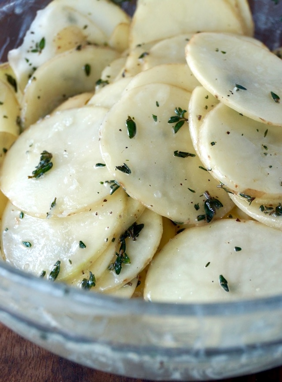 Glass bowl with thinly sliced potatoes coated with butter and fresh thyme.