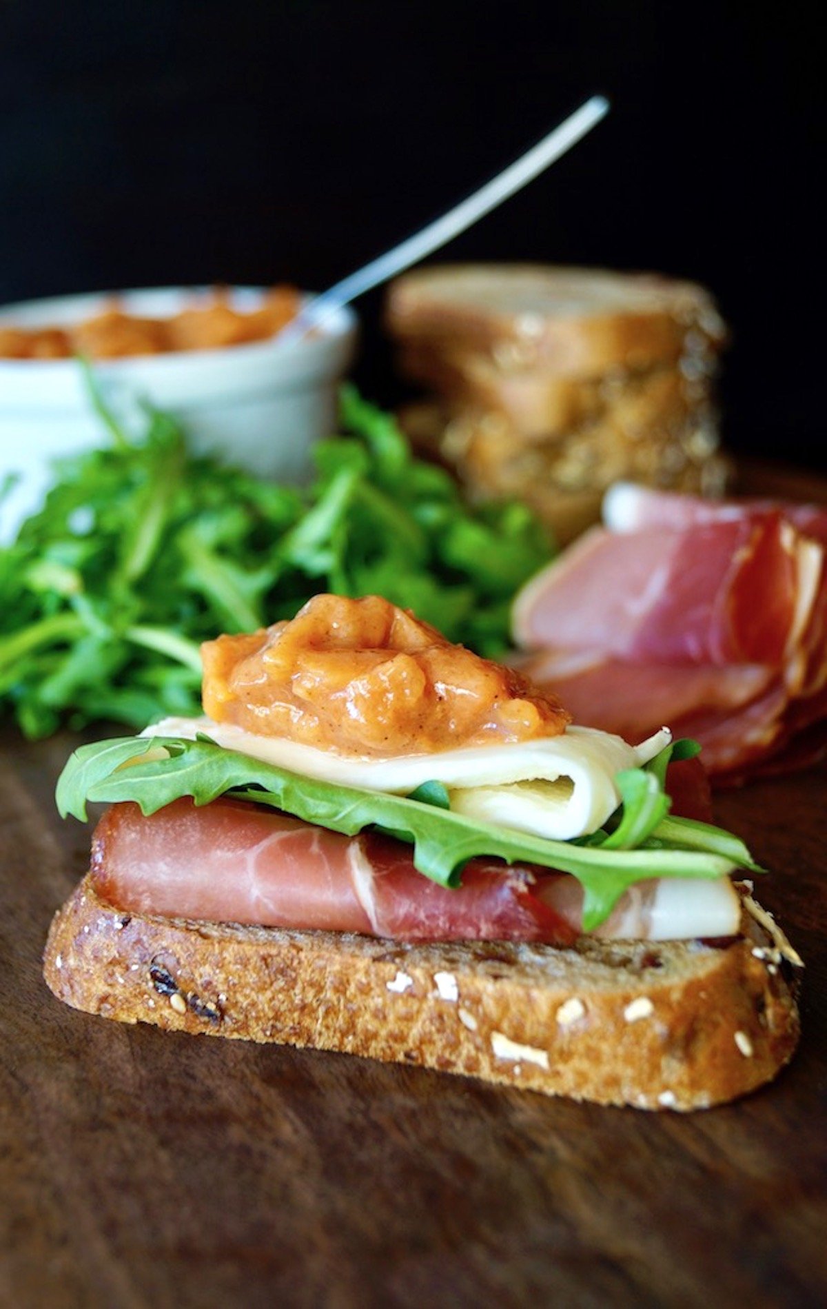 Open faced sandwich with Spicy Persimmon Preserves in a white bolw surrounded by meats, cheeses and bread slices.