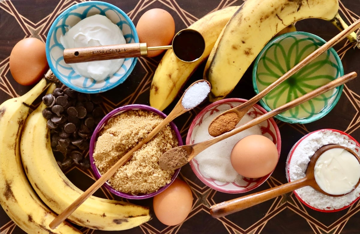 Ingredients for gluten-free banana cake including brown and white sugar, bananas, spices, sour cream, eggs, oil and vanilla - on a beautiful mixed wood cutting board.