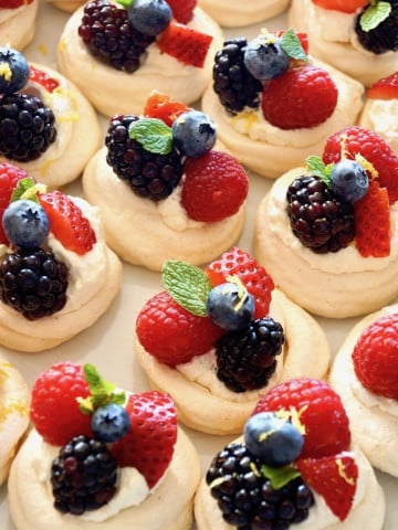 Several mini pavlova nests filled with fresh berries, whipped cream and a mint leaf, on a baking sheet.