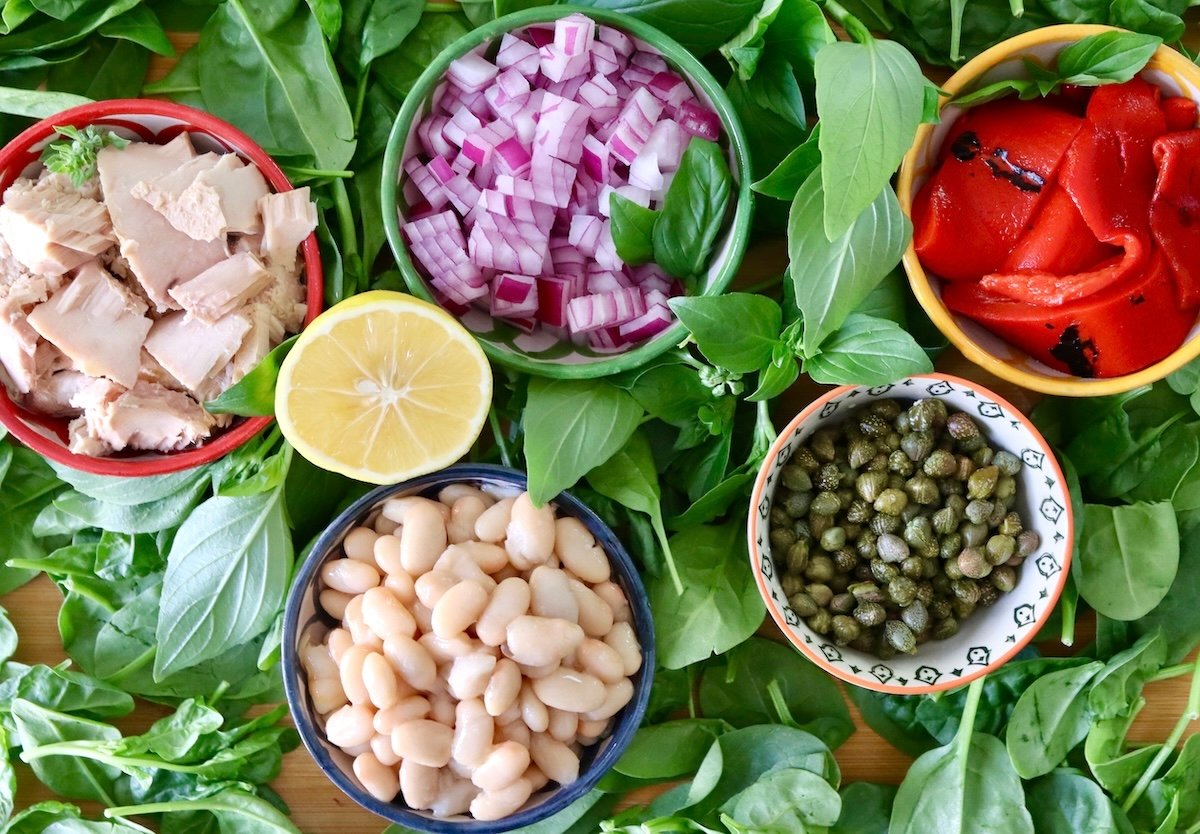 Ingredients for white bean tuna salad including red onion, white beans, capers, lemon, tuna  -- each in a small bowls surrounded by basil and spinach.