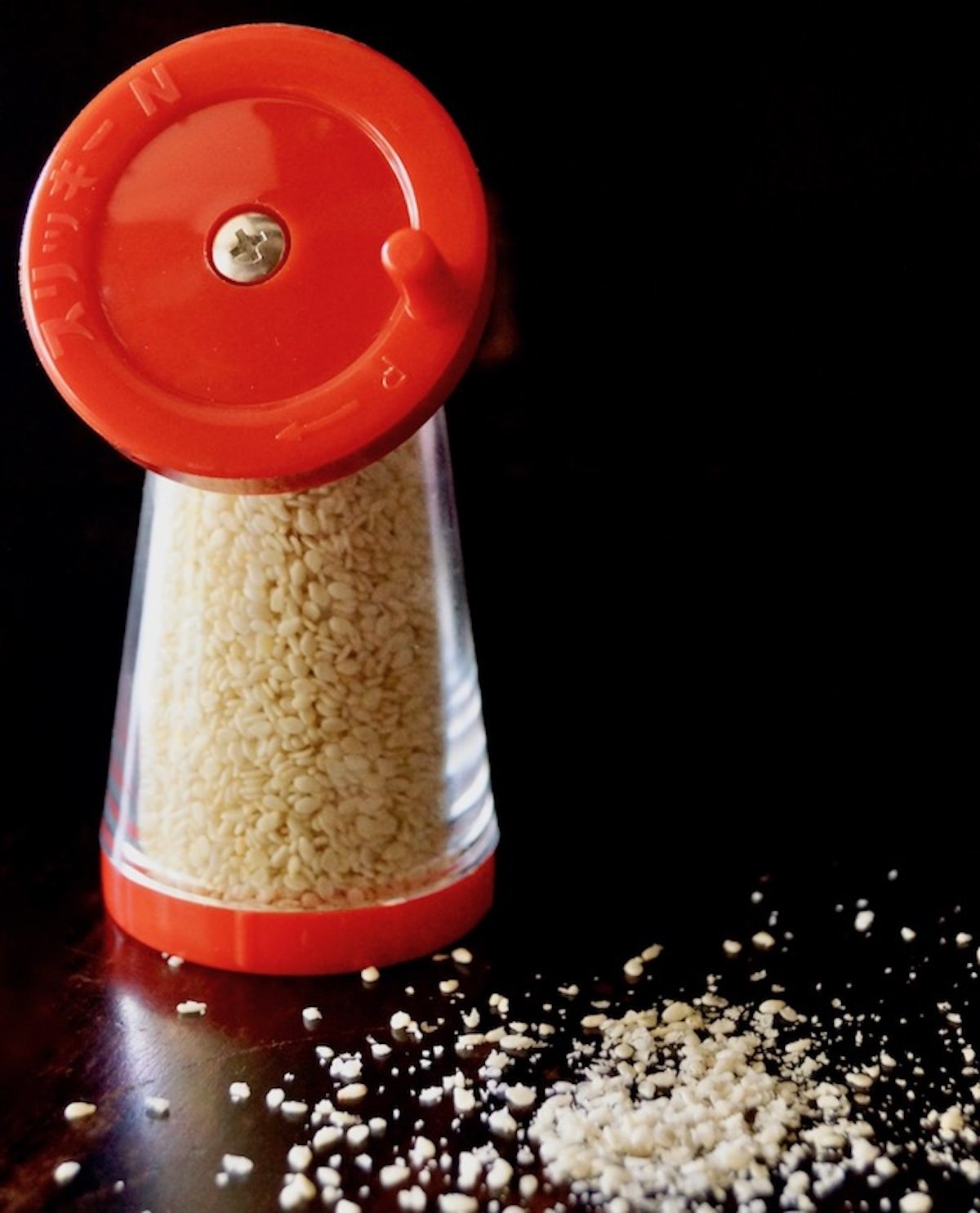 Red sesame seed grinder on black background with ground seeds next to it.