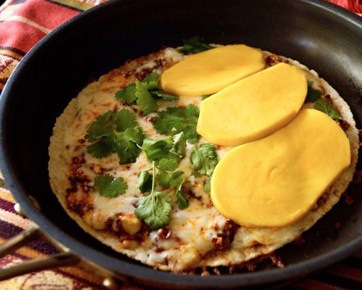 Tortilla in a pan with crumbled cheese mixed with red chili and mozzarella, cilantro and big mango slices.