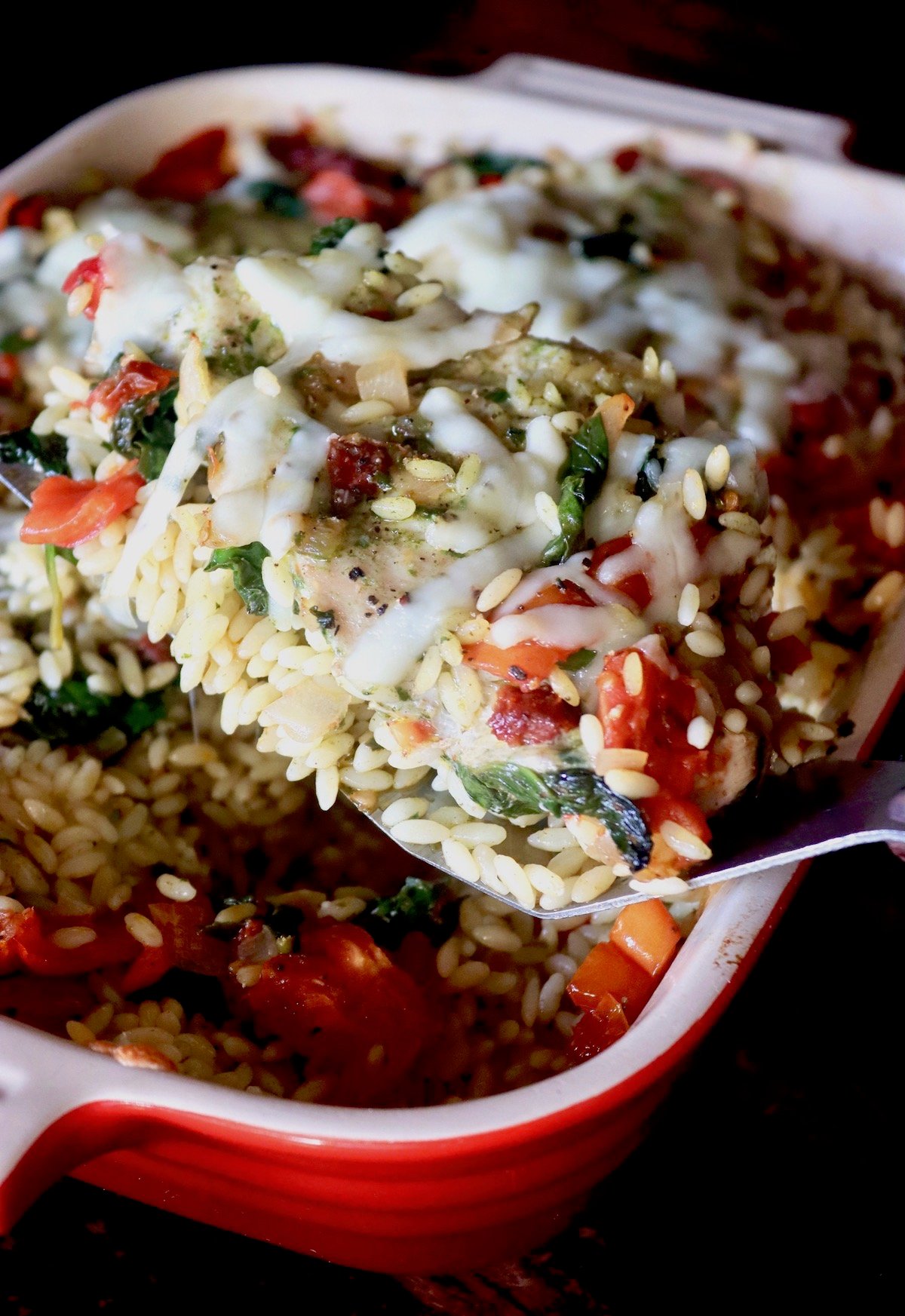 Red baking dish with orzo, chicken and tomatoes, with some of it lifted up on a spatula.