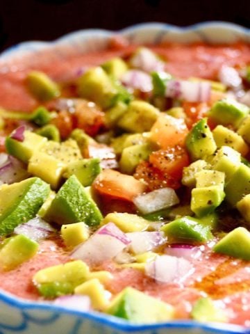 Close up of Easy Tomato Gazpacho recipe in a blue and white bowl with diced avocados, tomatoes and onion on top.