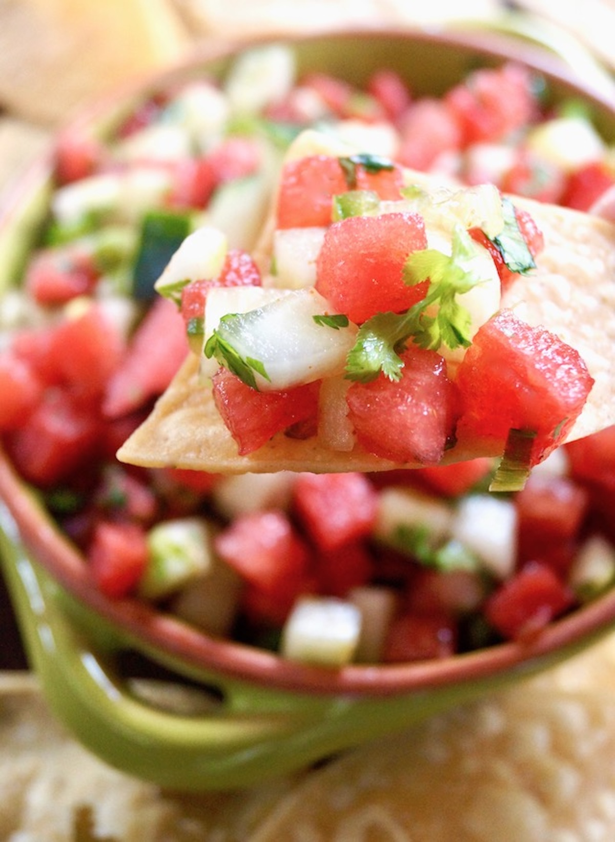 Watermelon cucumber salsa on a tortilla chip above above a bowl of the salsa.