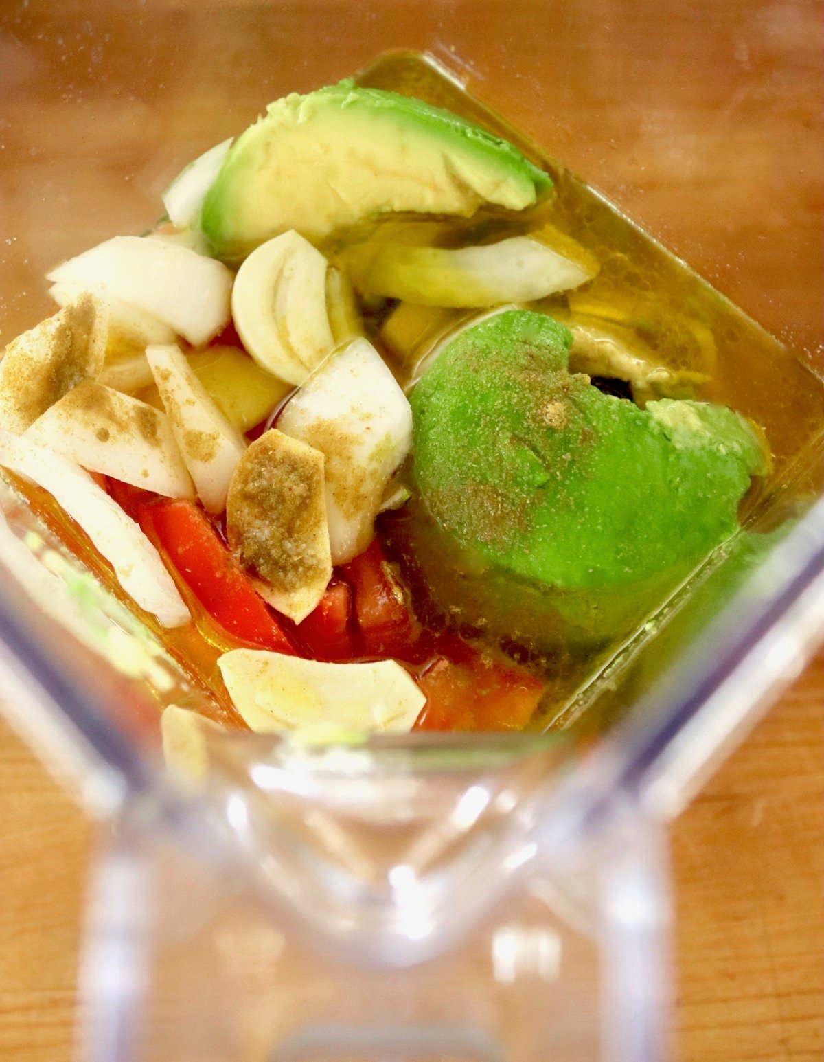 Blender filled with pieces of onion, avocado, spices, garlic and tomato.