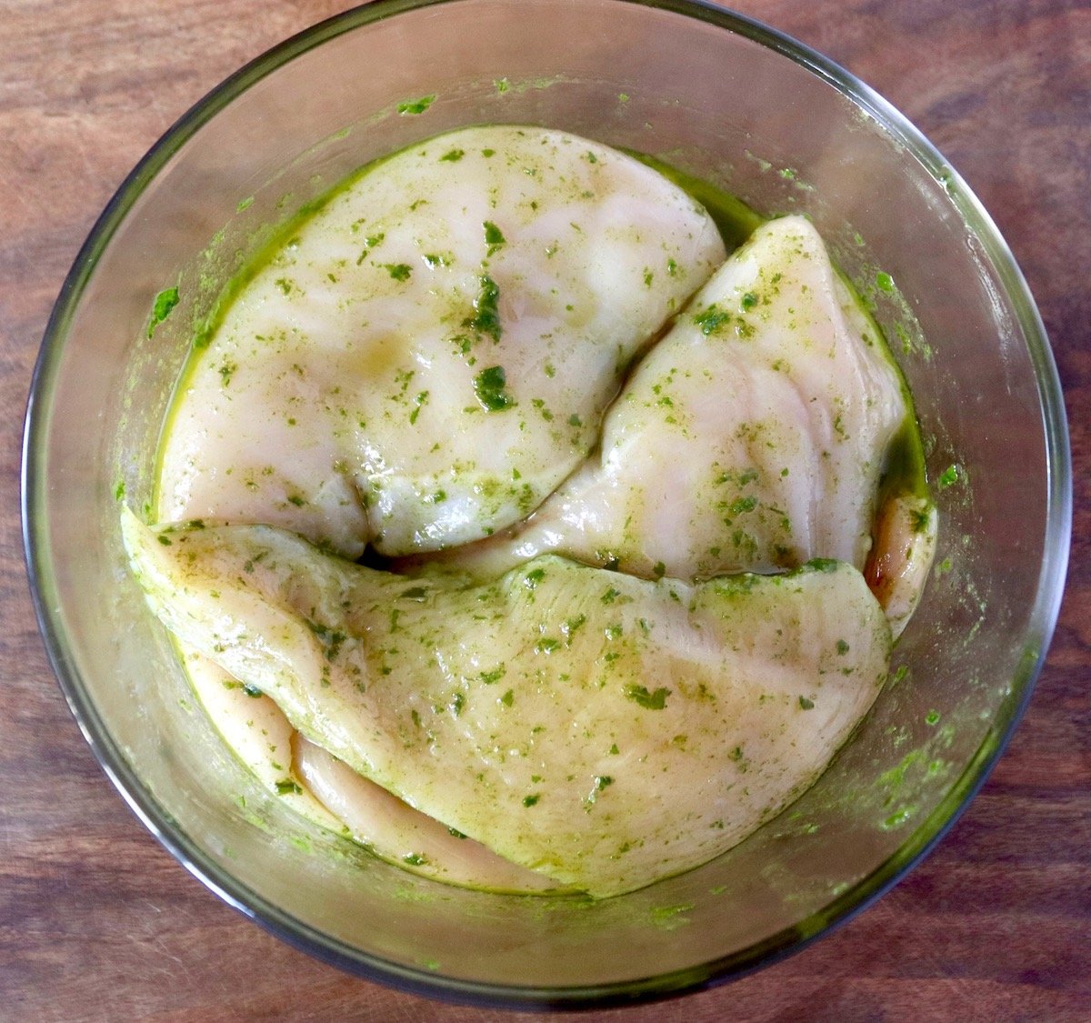 Three raw chicken breasts marinating in basil oil in a glass bowl.