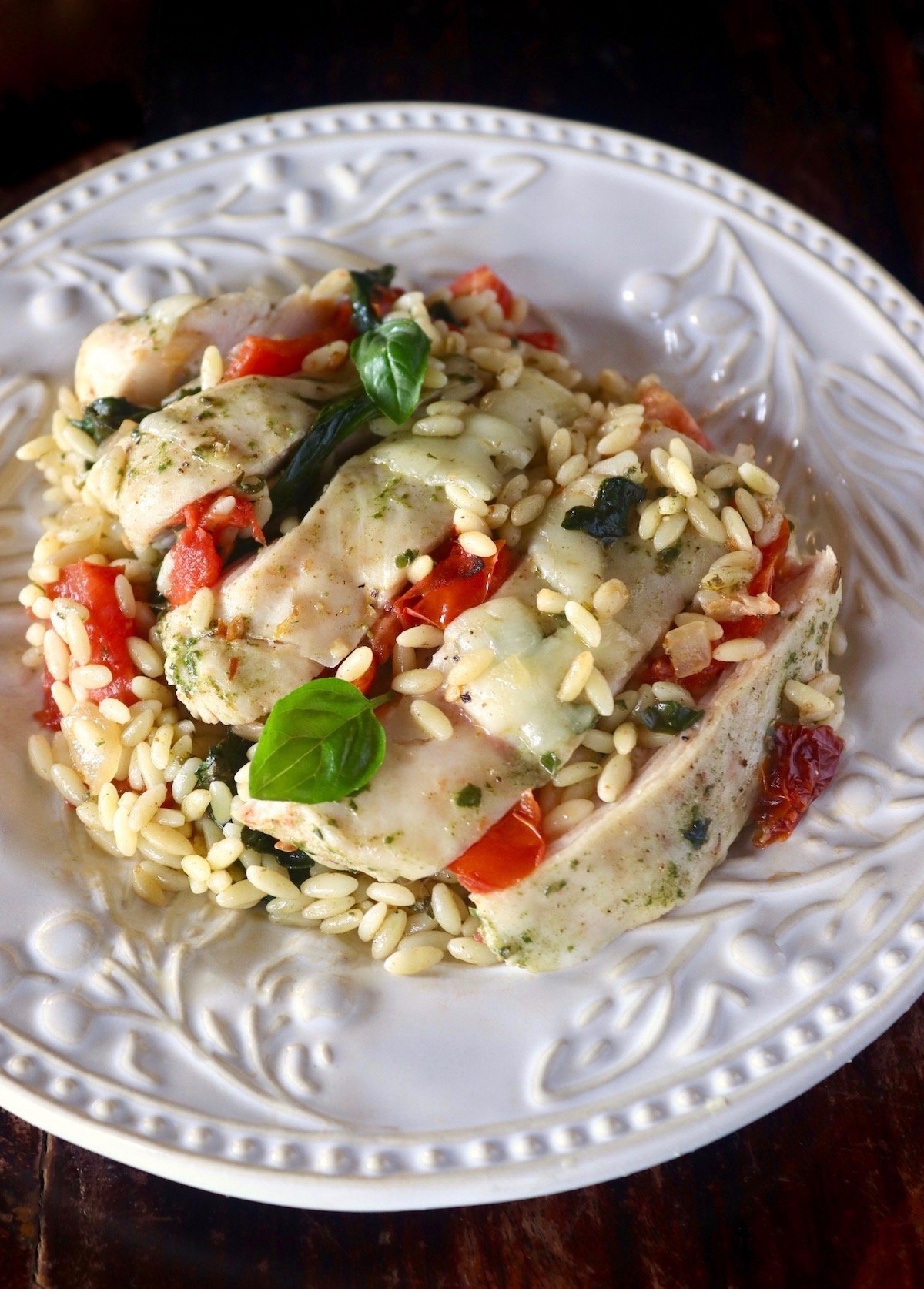 White plate with sliced Caprese chicken, basil, orzo and tomatoes.