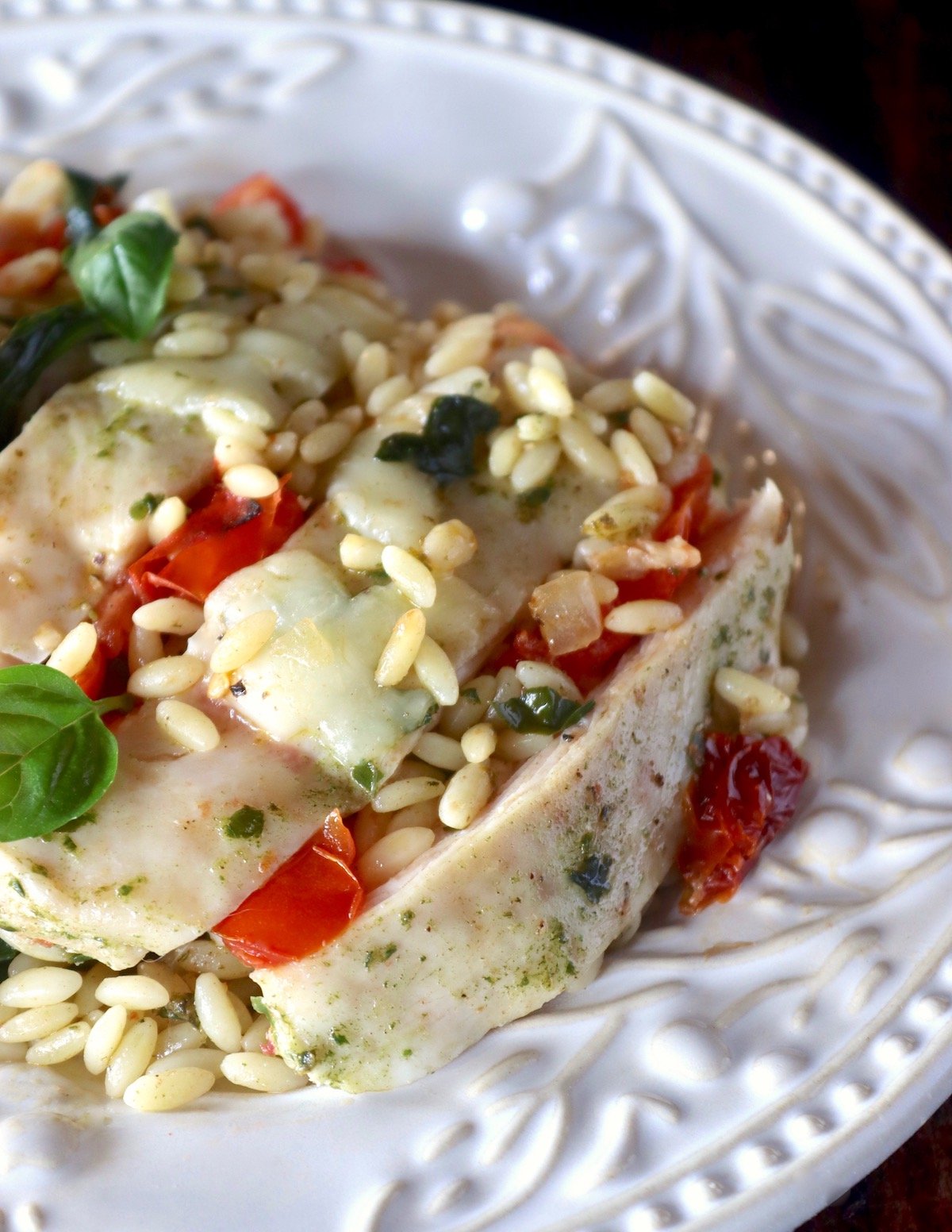 White plate with sliced chicken, basil, orzo and tomatoes.
