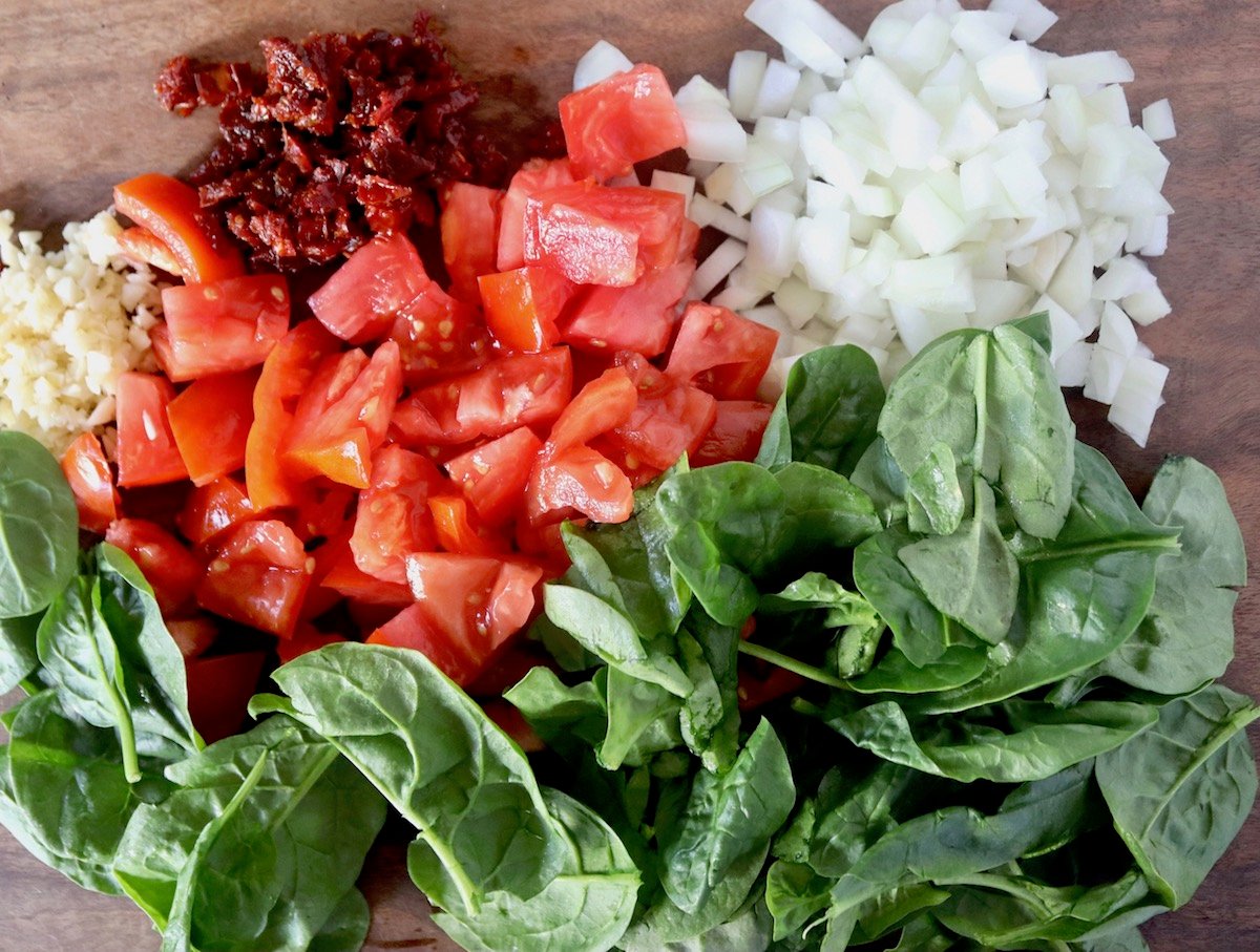 Spinach leaves, chopped tomatoes, onion, garlic and sun-dried tomatoes on a wood board.