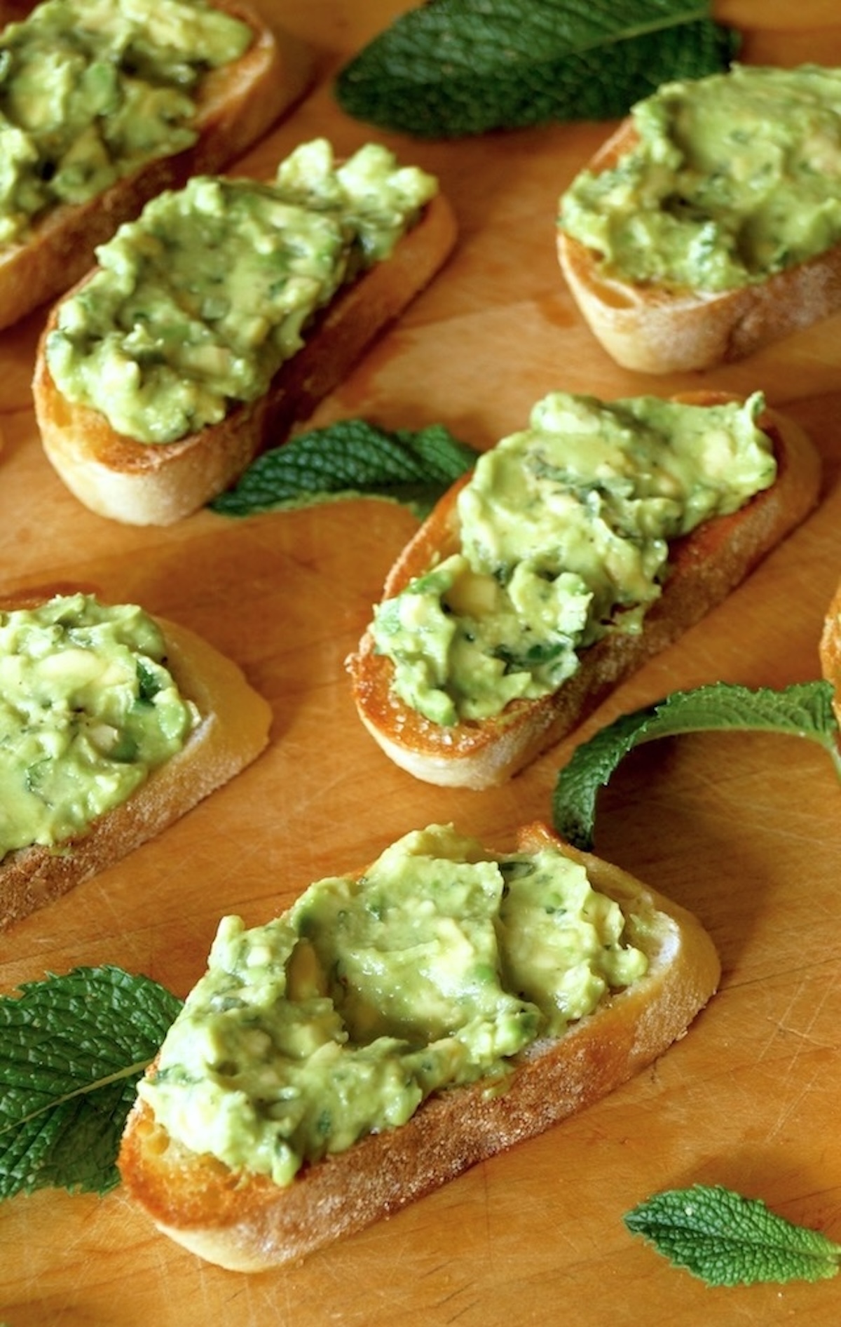 Several small slices of avocado toast with fresh mint on a wooden cutting board.