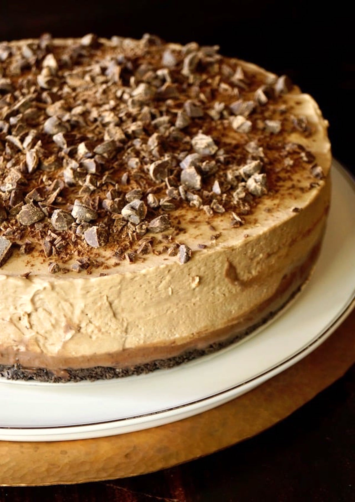 No-Bake Espresso Cheesecake with Ganache and a layer of chopped chocolate on top, on a white cake stand.