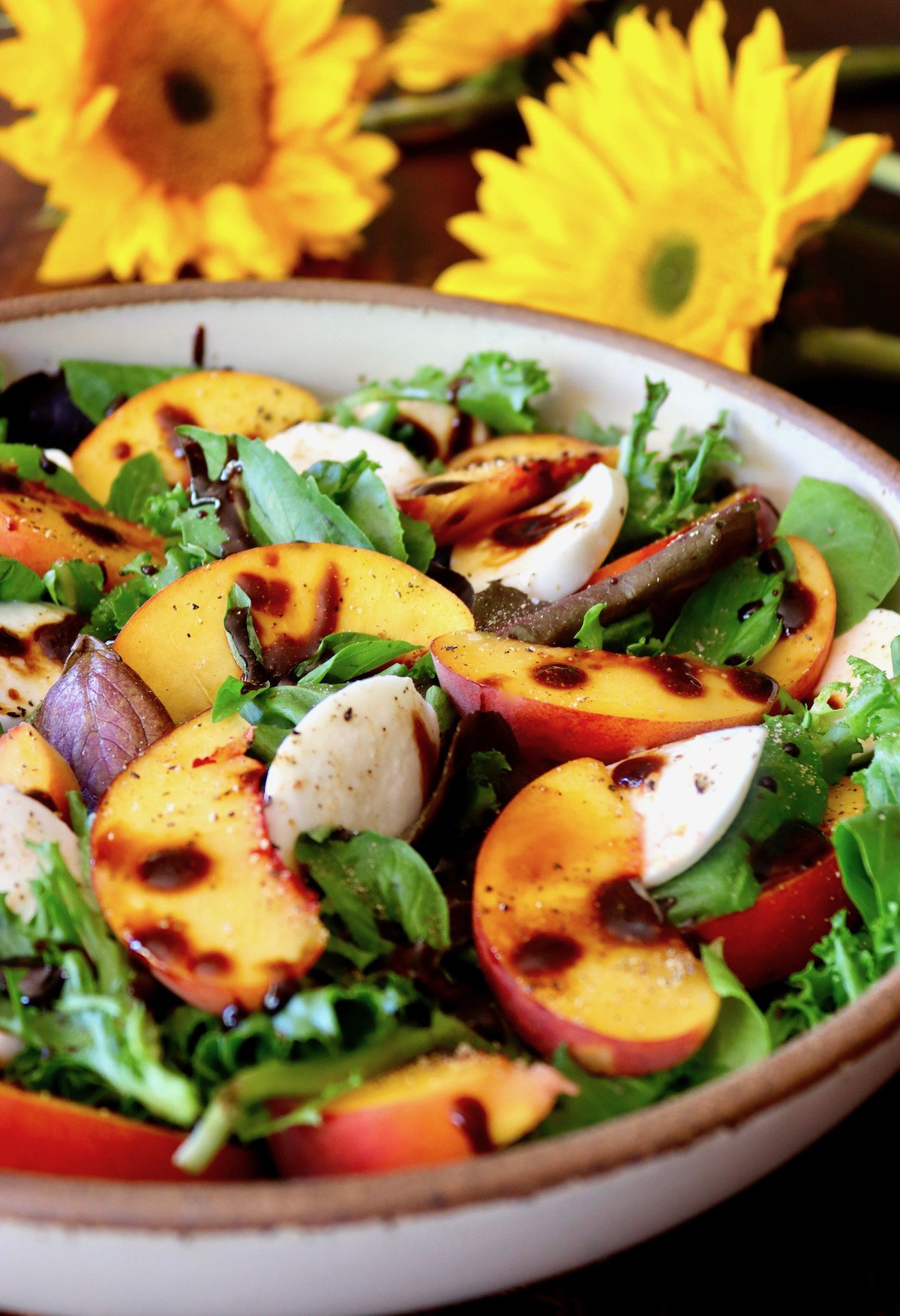 Cream-colored ceramic bowl filled to the top with Peach Caprese Salad with drops of balsamic glaze and fresh sun flowers behind it.